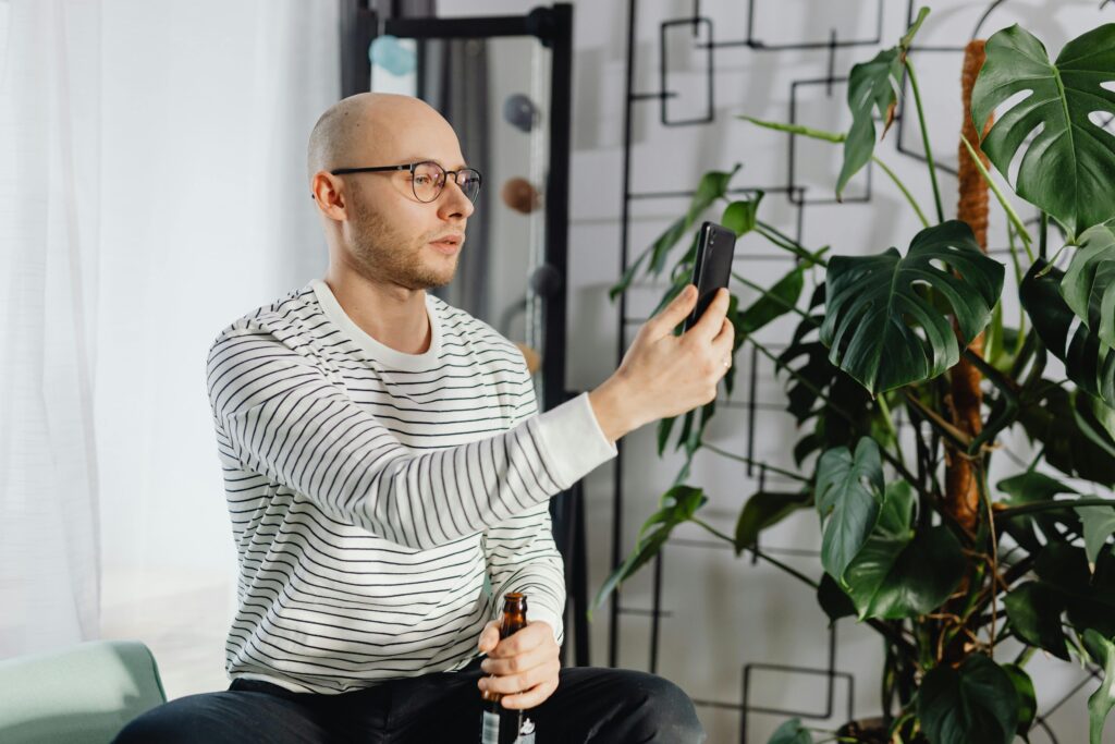 ambiente de sala bem iluminado, com um homem de oculos olhando para o celular, ao lado planta grande de costela-de-adao.