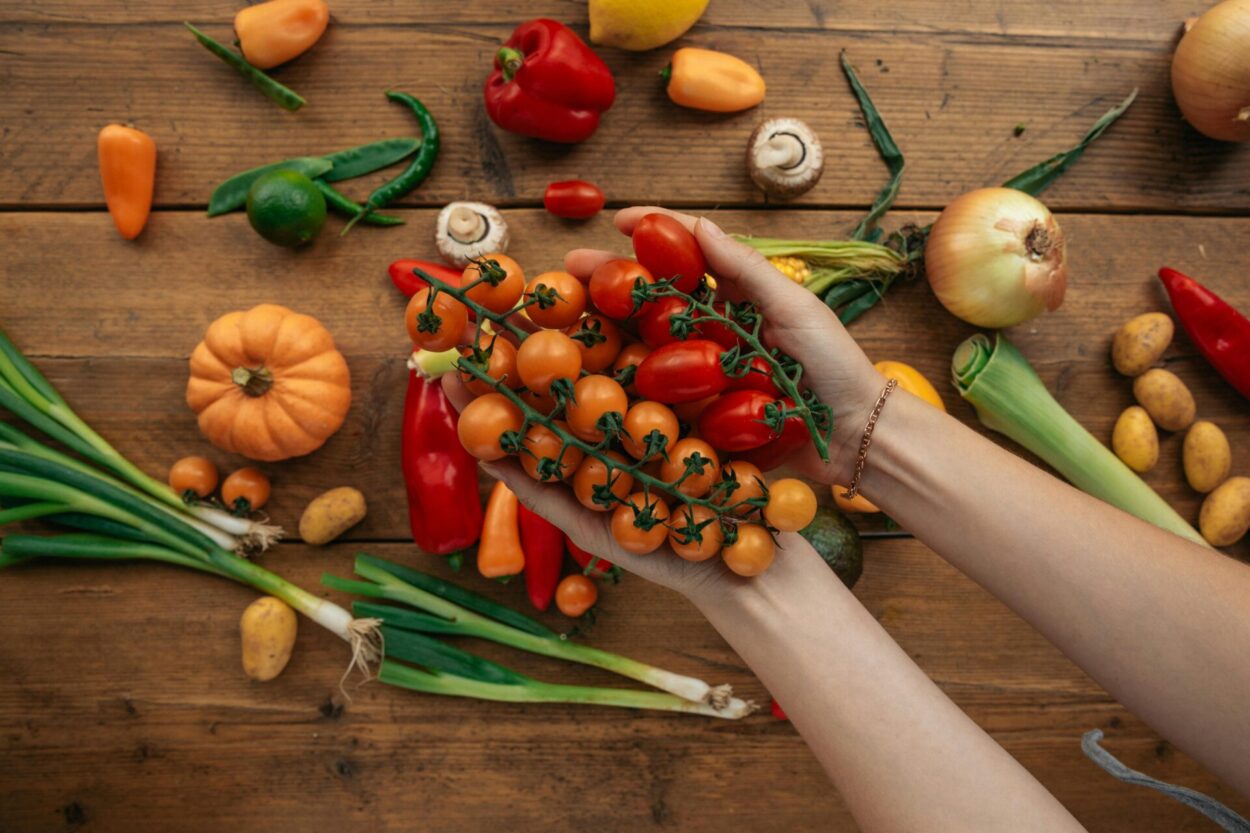 mãos em cima de bancada de madeira segurando tomatinhos, cebolinha e na bancada diversos como abobora, pimentao vermelho, pimentao amarelo, cebolinha
