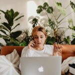 ambiente de sala com mulher trabalhando em laptop e ao fundo diversas plantas de flohagem grande.